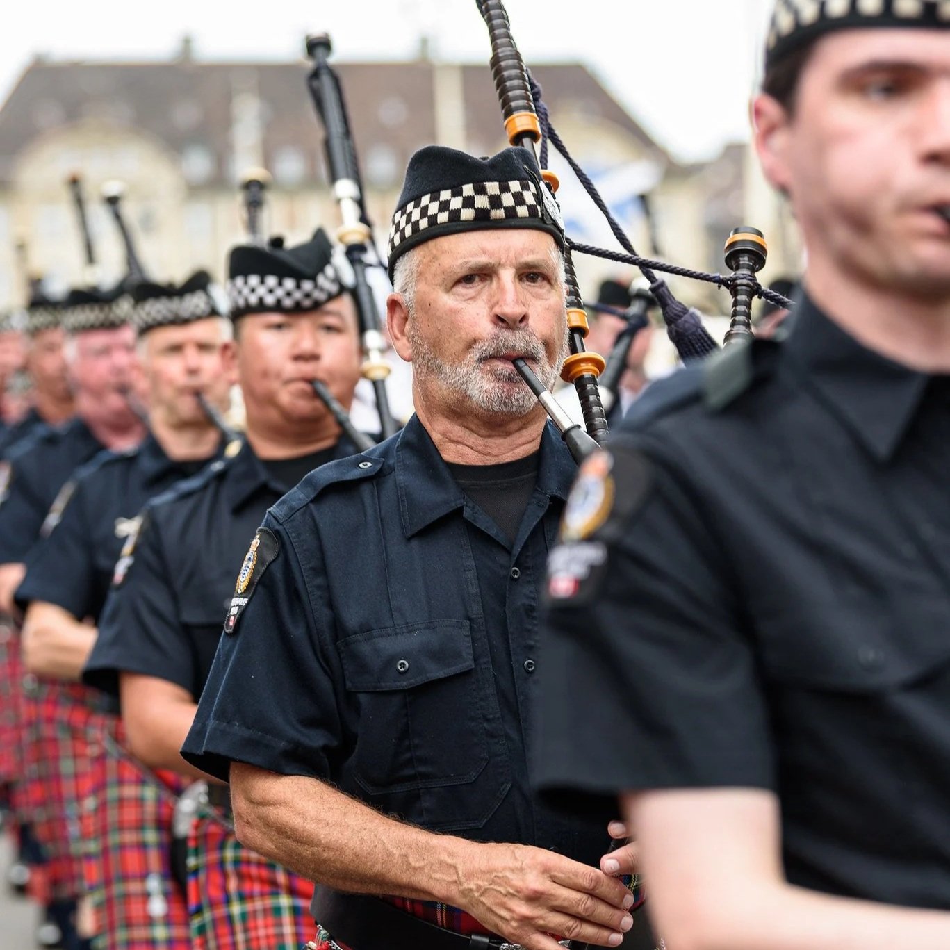 A line of police officers playing bagpipes during a parade, dressed in navy uniforms and Scottish kilts.