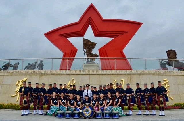 Group of people in traditional attire posing in front of a large red star monument with a golden eagle sculpture, cloudy sky background.