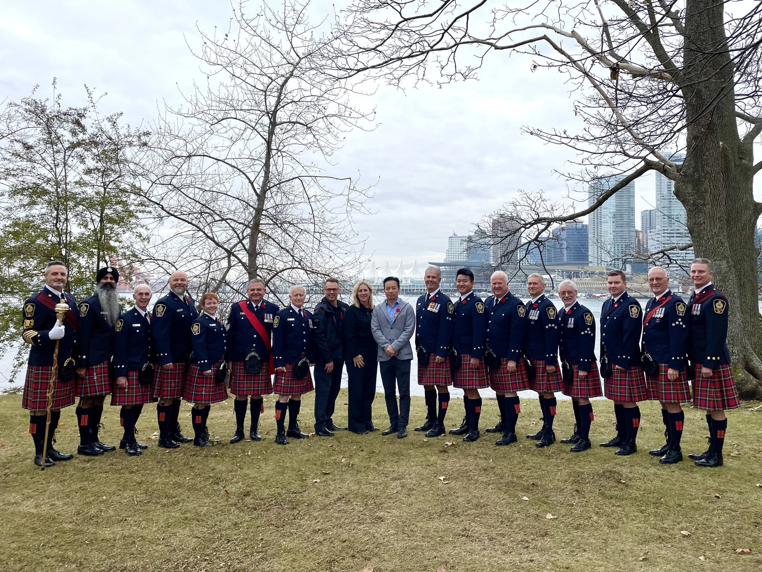 A group of people dressed in formal Scottish kilts and police uniforms standing outdoors on grass with leafless trees, city skyline, and water in the background.