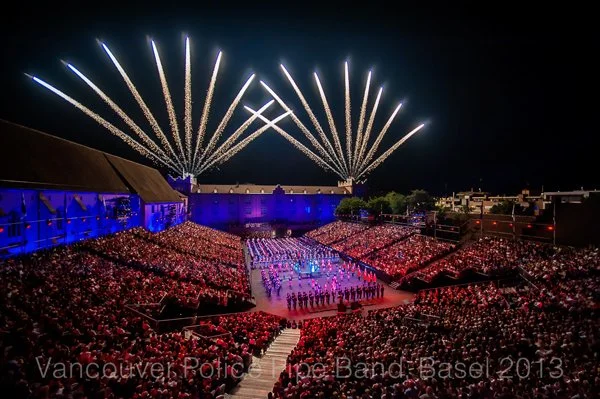 A large outdoor concert with a crowd seated in front of a stage, fireworks lighting up the night sky.