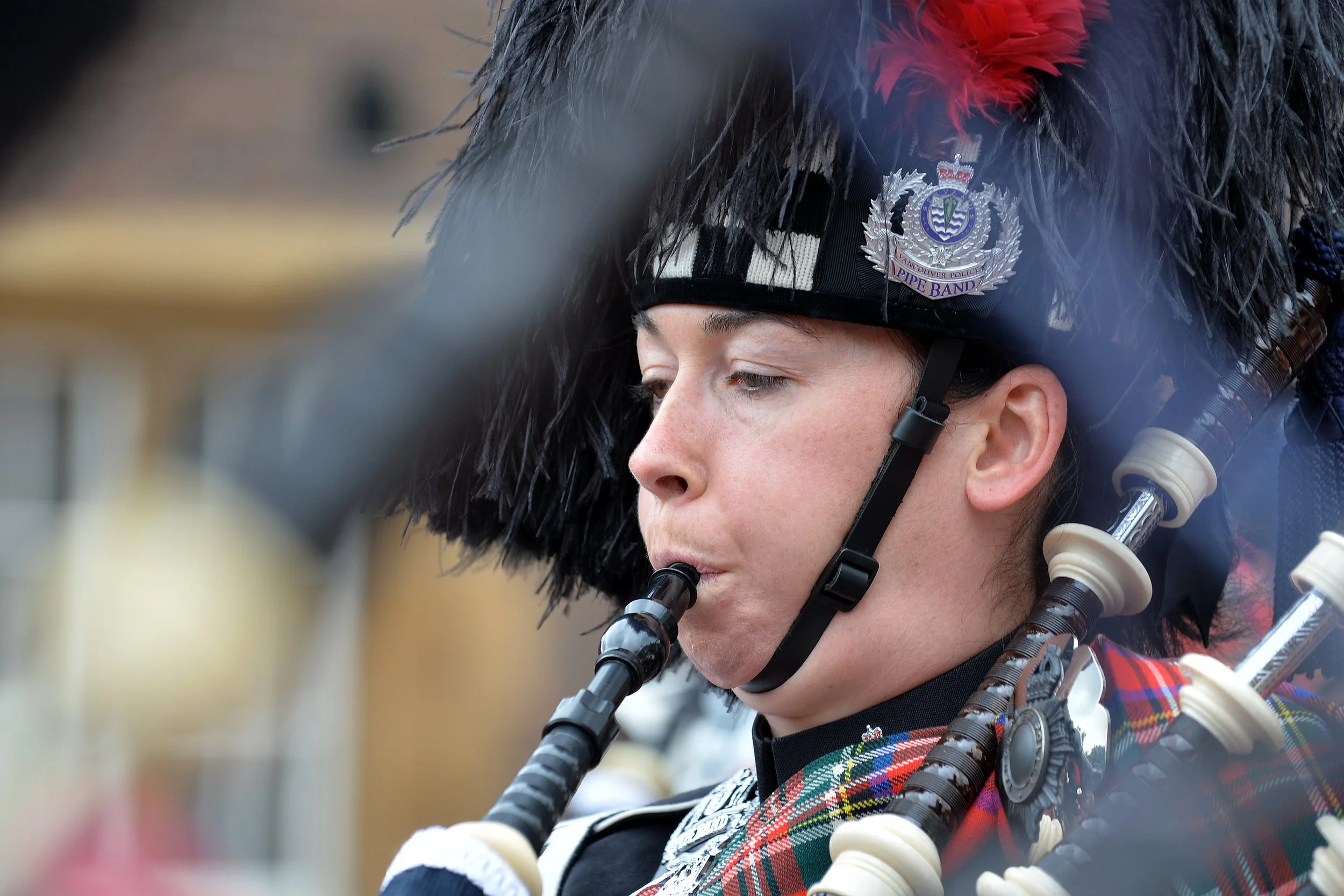 A woman playing the bagpipes, wearing a Black Watch tartan uniform and a feathered hat with a Police Band badge.