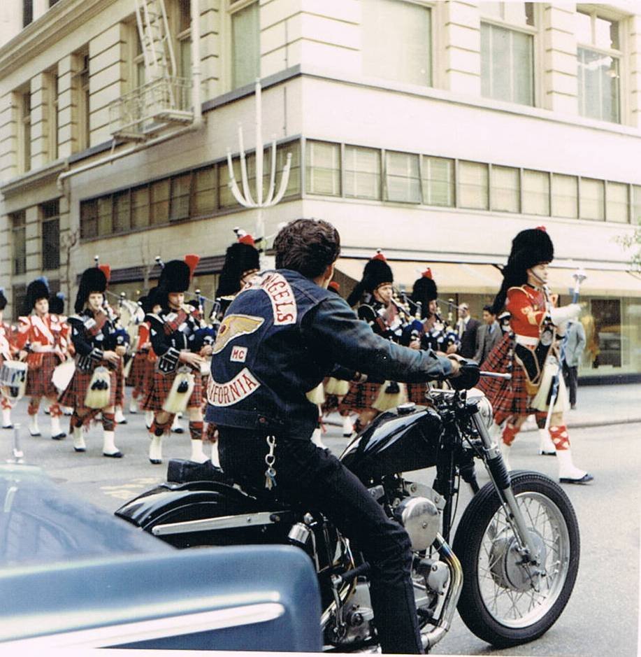 A motorcyclist wearing a black jacket riding past a parade with marching band members dressed in Scottish attire, including kilts and feathered hats, on a city street.