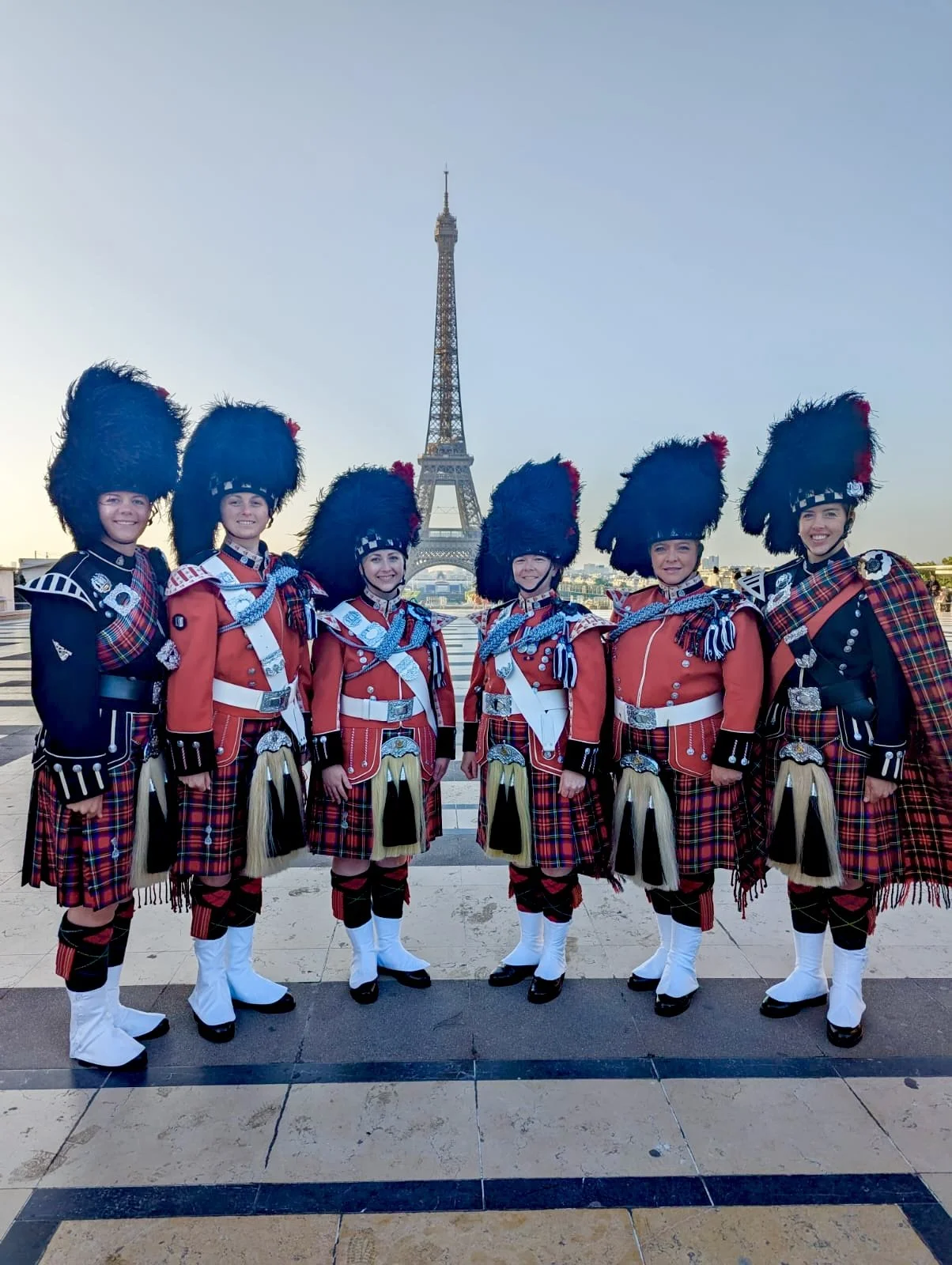 Six women dressed in traditional Scottish Highland dress with the Eiffel Tower in the background, standing on a terrace.