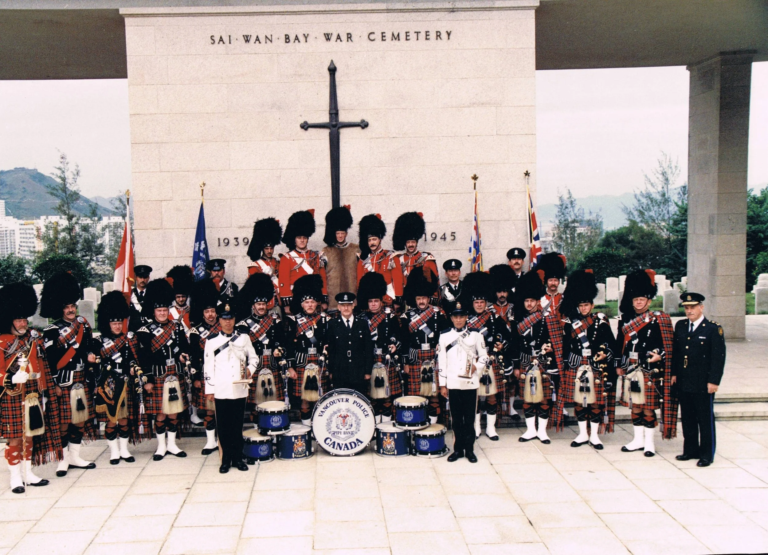 Group of people in traditional Scottish attire, including kilts and bearskin hats, posing in front of a war memorial at Sai Wan Bay War Cemetery, with flags and a drum labeled Vancouver Police.
