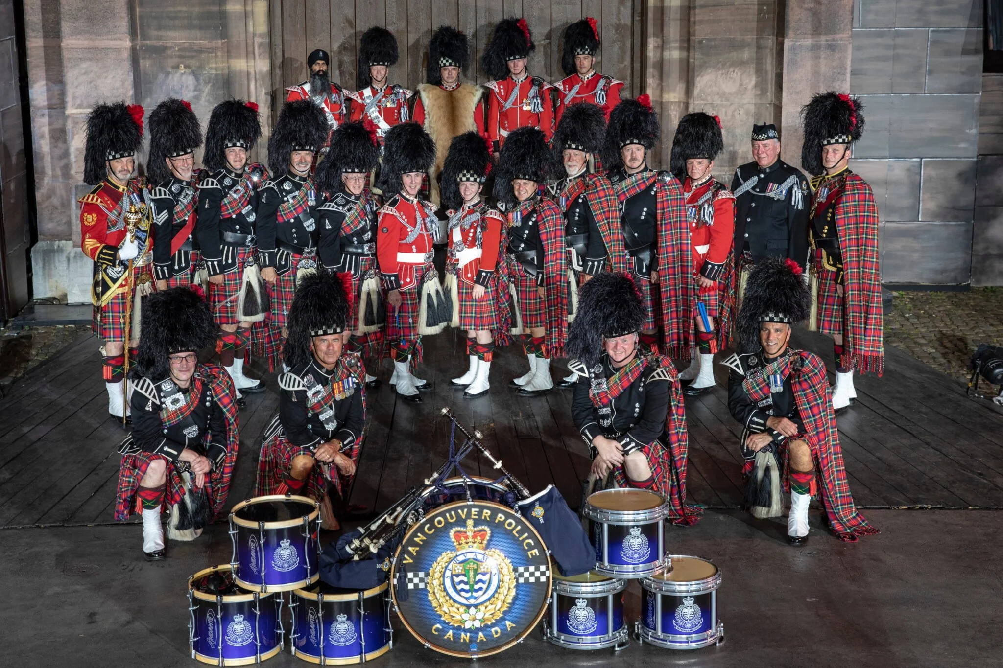 A group of people dressed in traditional Scottish Highland dress, including kilts, sporrans, and feathered hats, posing in front of a building. Some are holding musical instruments with a Vancouver Police badge on the drums visible in the foreground.