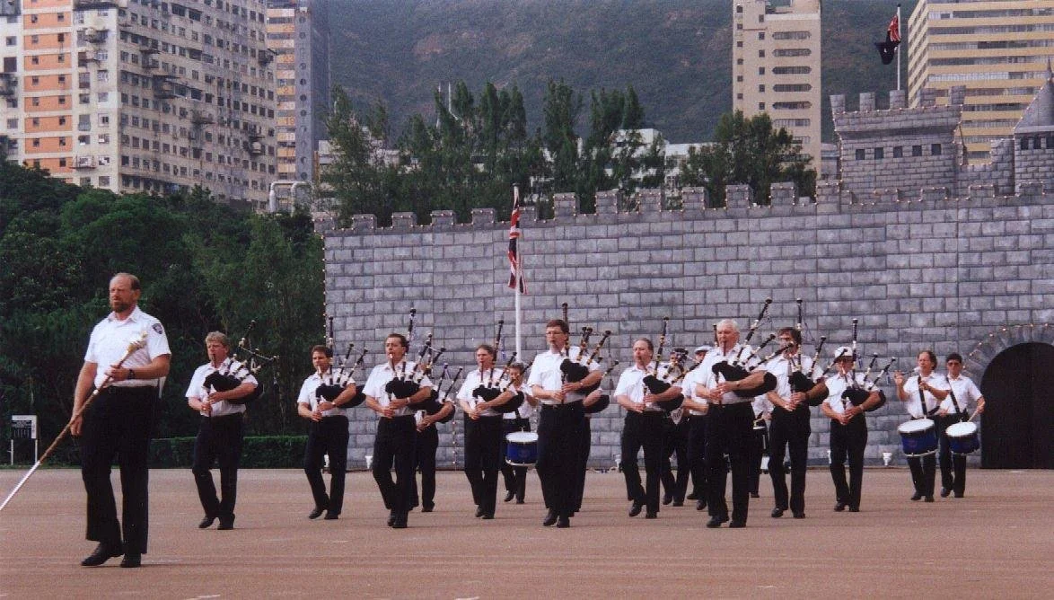 A group of uniformed musicians playing bagpipes and drums outdoors in front of a castle-like structure, with modern city buildings in the background.