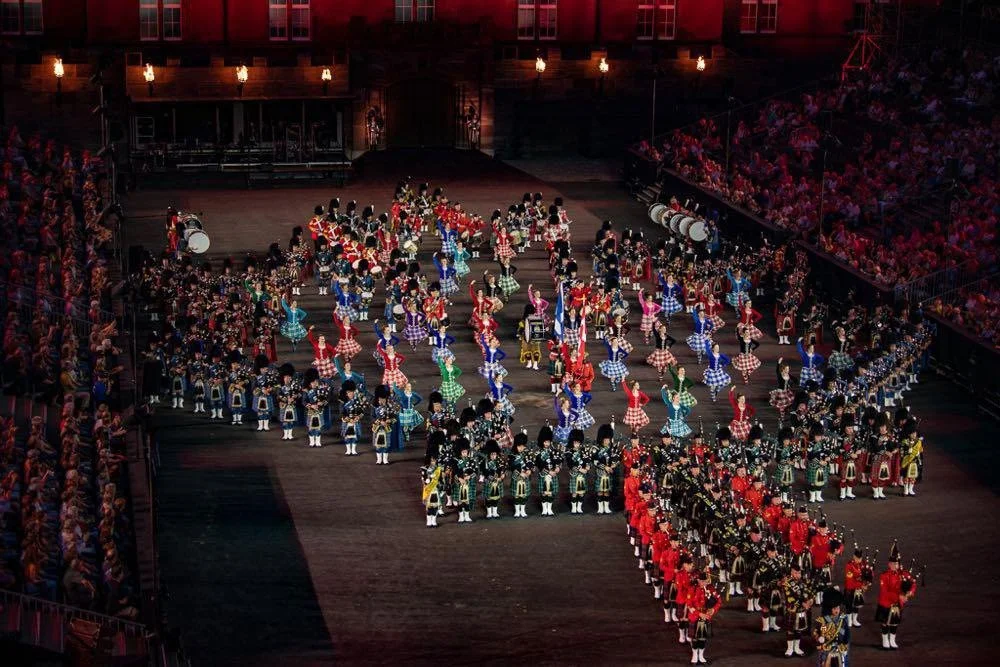 A large group of colorfully dressed dancers performing on a stage, with an audience on either side, in a theatrical setting.