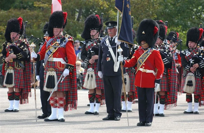 A group of people in traditional Scottish military uniforms, including kilts and feathered hats, participating in a parade or ceremony outdoors.