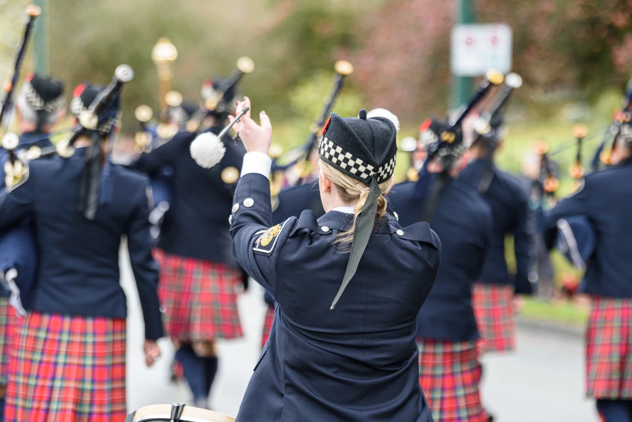 A woman in a uniform leading a marching band, all wearing dark jackets with checked plaid skirts, during an outdoor parade or event.
