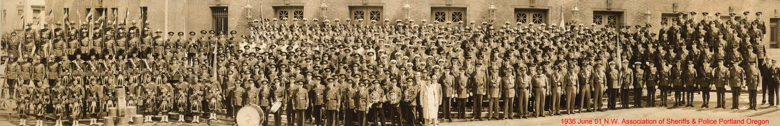 A large group of police officers and sheriffs in uniform posing for a photo in front of a brick building in June 1936, with some officers holding flags and drums, and a person dressed in white standing in front center.