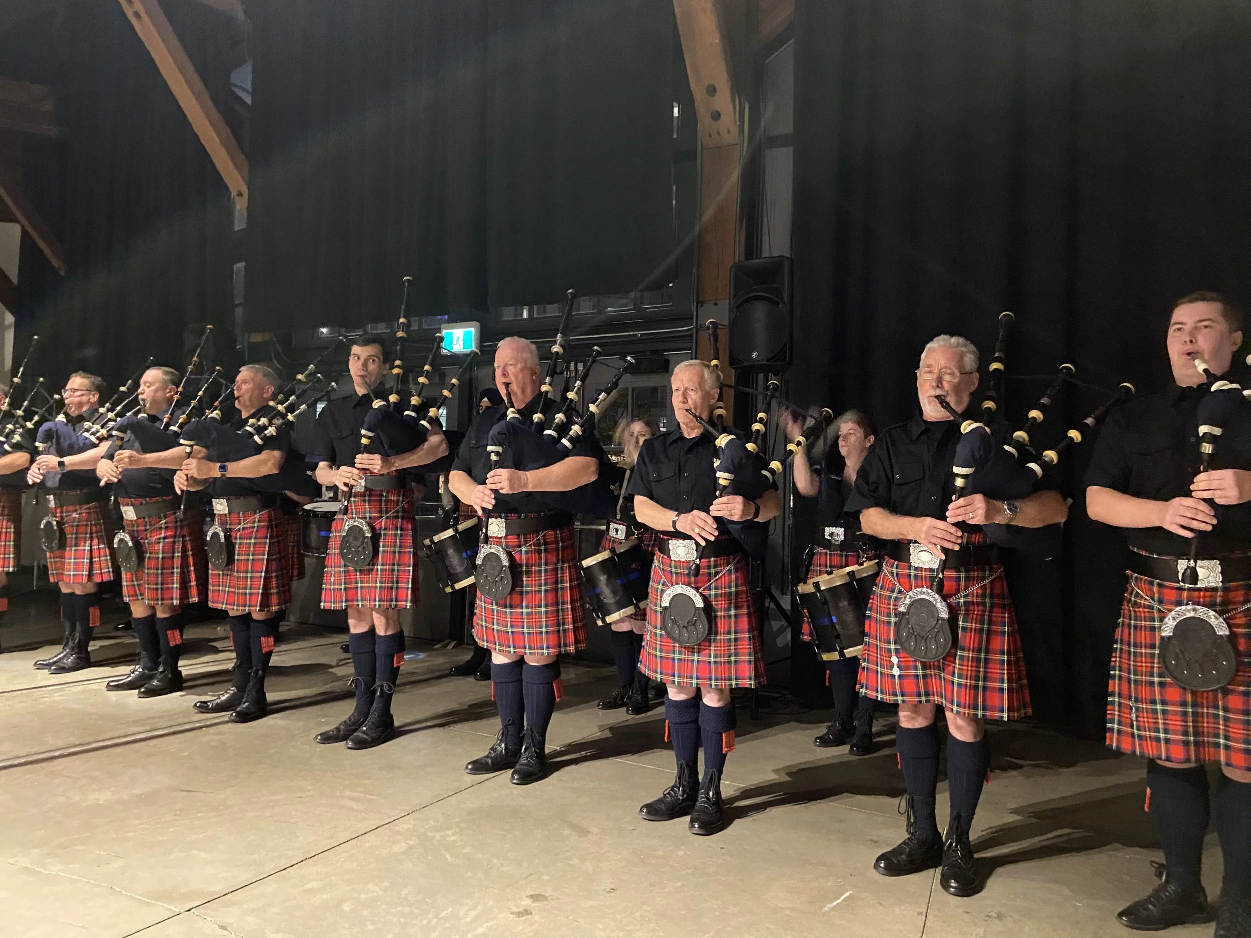 A group of men dressed in traditional Scottish attire, wearing kilts and black shirts, playing bagpipes on a stage.