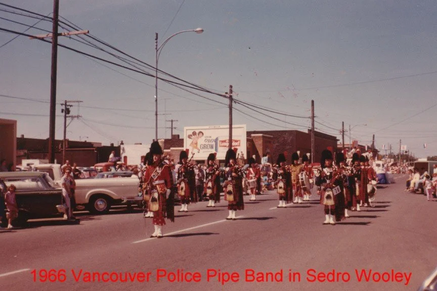 A parade featuring the Vancouver Police Pipe Band in Sedro Woolley in 1966, with band members dressed in traditional Scottish uniforms marching on a city street, lined with vintage cars and onlookers.