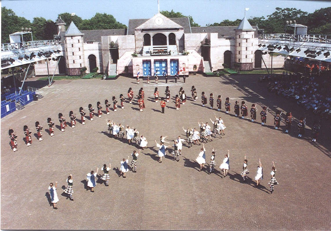 Children performing a dance in front of a castle-shaped stage, dressed in traditional Scottish attire including kilts and dresses, with an audience watching from the sides.