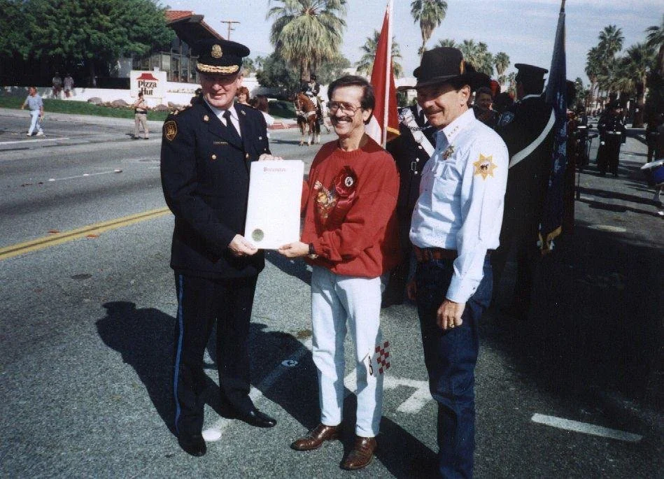 Three men standing on a street during a parade, with two of them in uniform and the third in casual clothing, smiling and holding a large envelope or certificate. Behind them, people are walking and horse-mounted parade participants are visible, with palm trees and a Pizza Hut sign in the background.