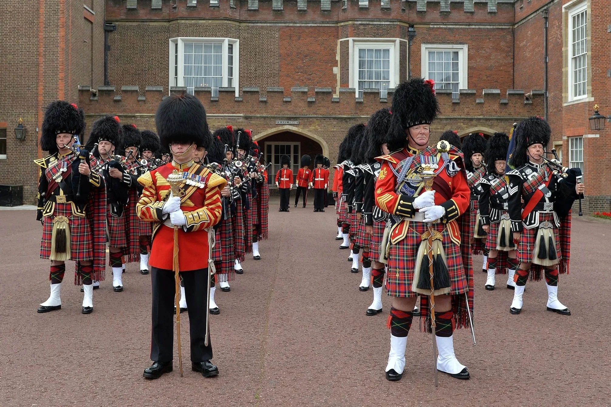 A parade of soldiers in traditional Scottish Highland dress, including kilts and bearskin hats, marching in front of a historic brick building.