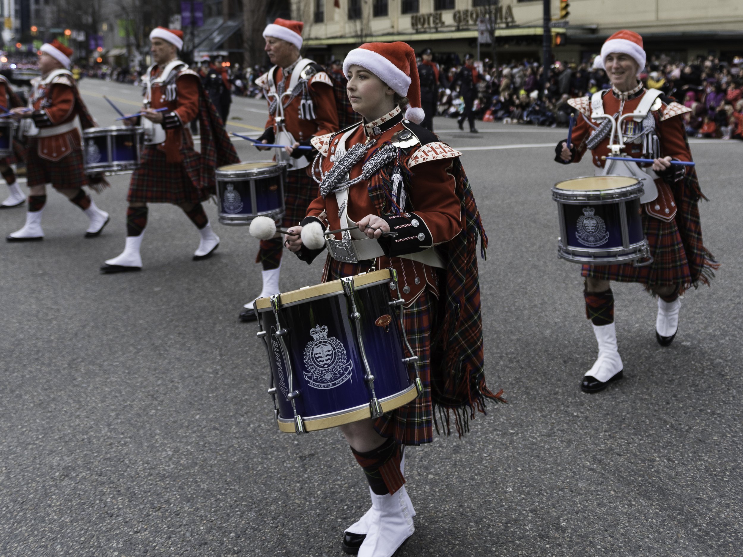 A group of parade drummers dressed in festive Scottish attire with tartan kilts and red jackets, marching down a city street during a holiday parade, with spectators lining the street in the background.