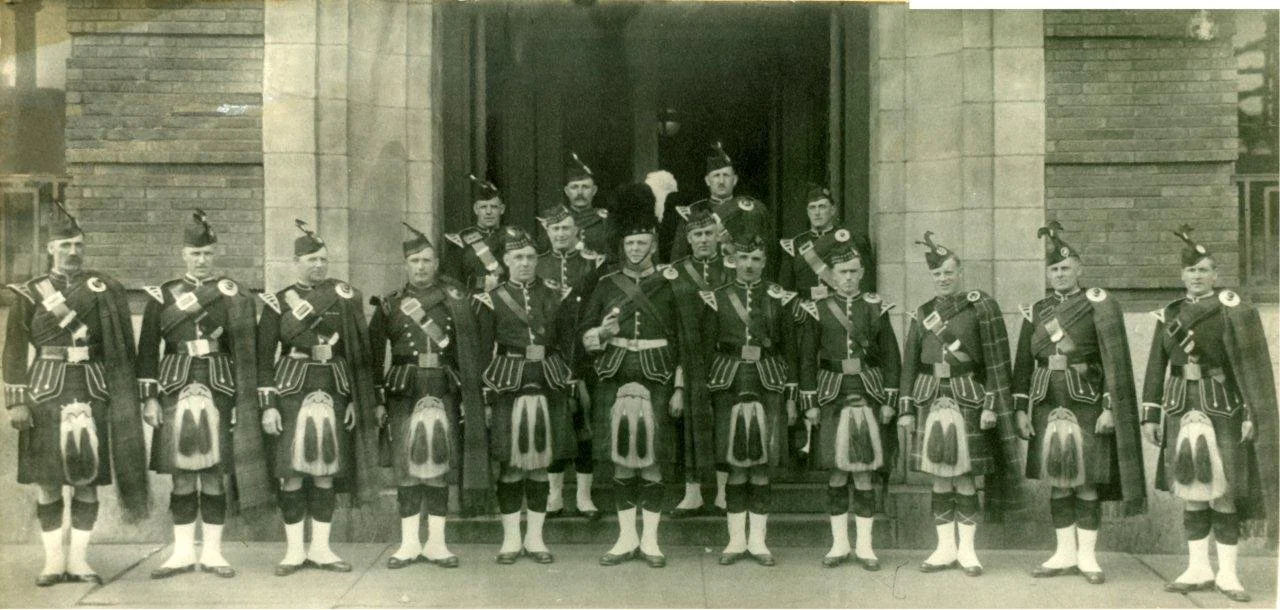 Group of men dressed in traditional Scottish Highland dress, including kilts, jackets, and feathered hats, standing in front of a stone building.