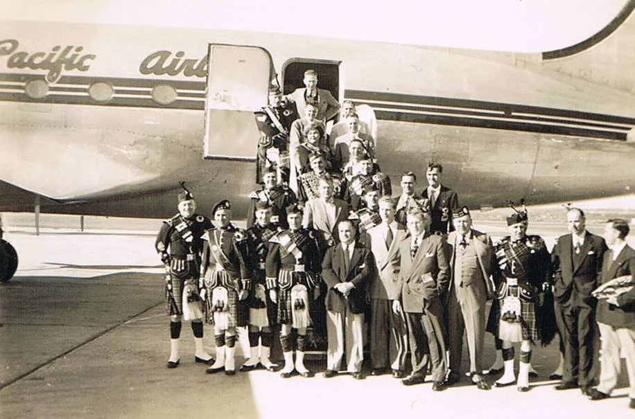 A group of people, including military personnel in traditional Scottish kilts, posing on an airport tarmac in front of a Pacific Air airplane. Some are standing on the stairs leading to the airplane's entrance, while others stand on the ground.