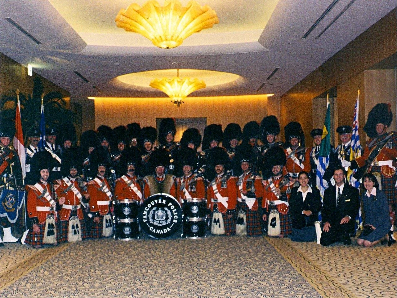Group of people dressed in Scottish traditional attire, including kilts and bearskin hats, posing for a photo in a conference room with chandeliers. The drums in the center have the text 'Vancouver Police Canada'.