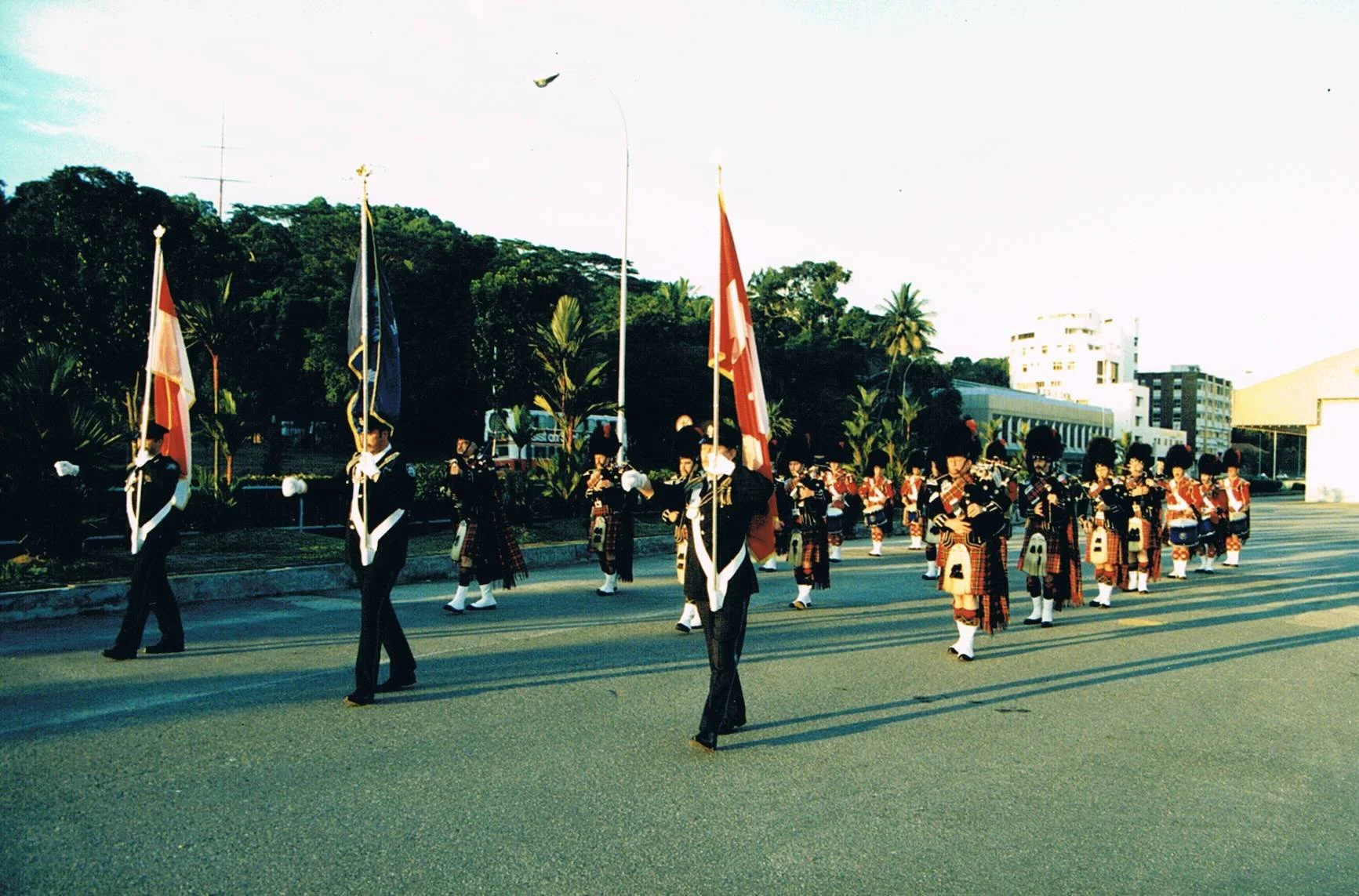 A parade with uniformed members marching and carrying flags, including the Canadian flag, in an outdoor urban setting with buildings and green trees in the background.
