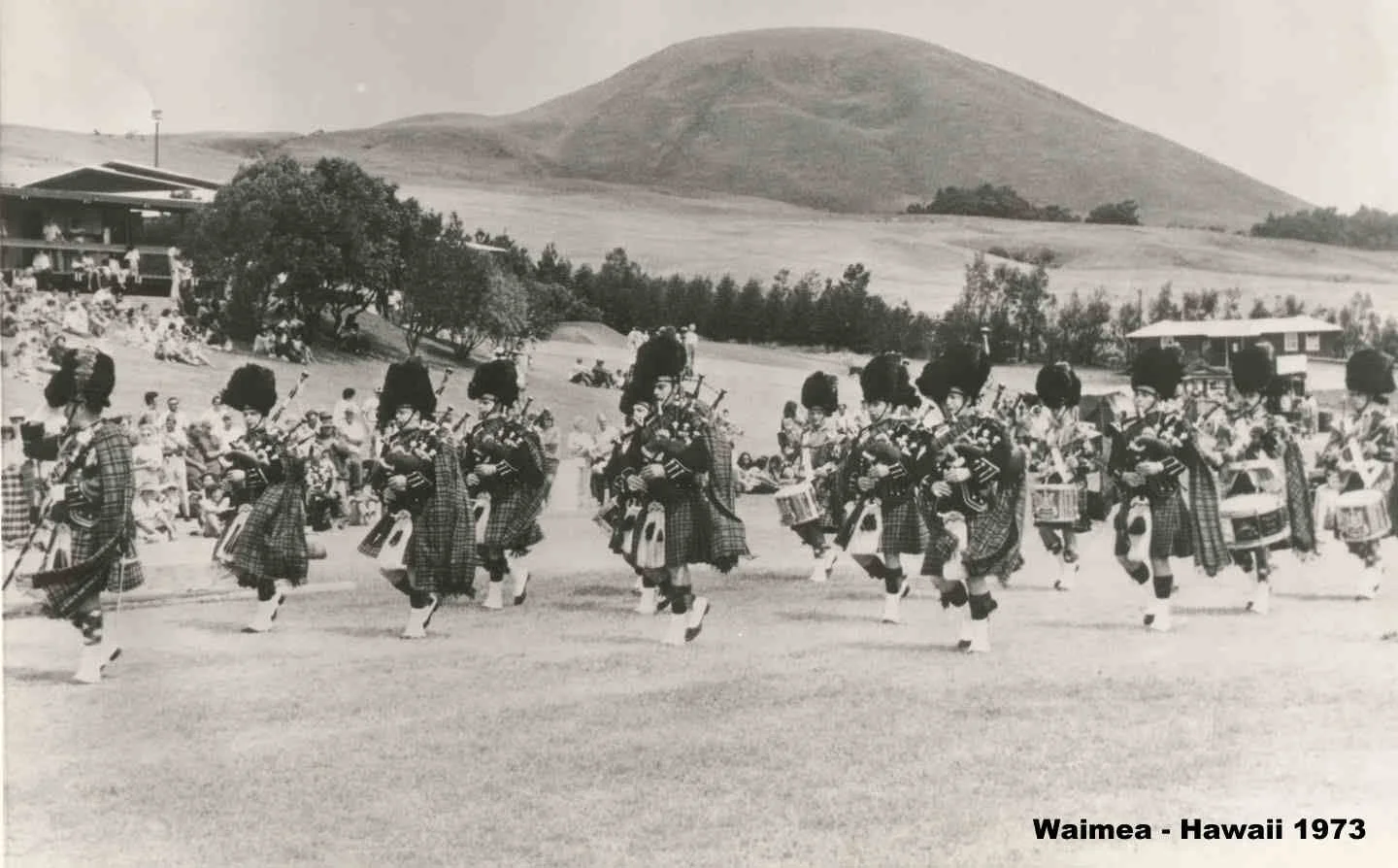 Black and white photograph of a traditional Hawaiian hula dance performance in Waimea, Hawaii, 1973. Several women in plaid skirts and headgear dance with musical instruments in an open field, with spectators and scenic hills in the background.