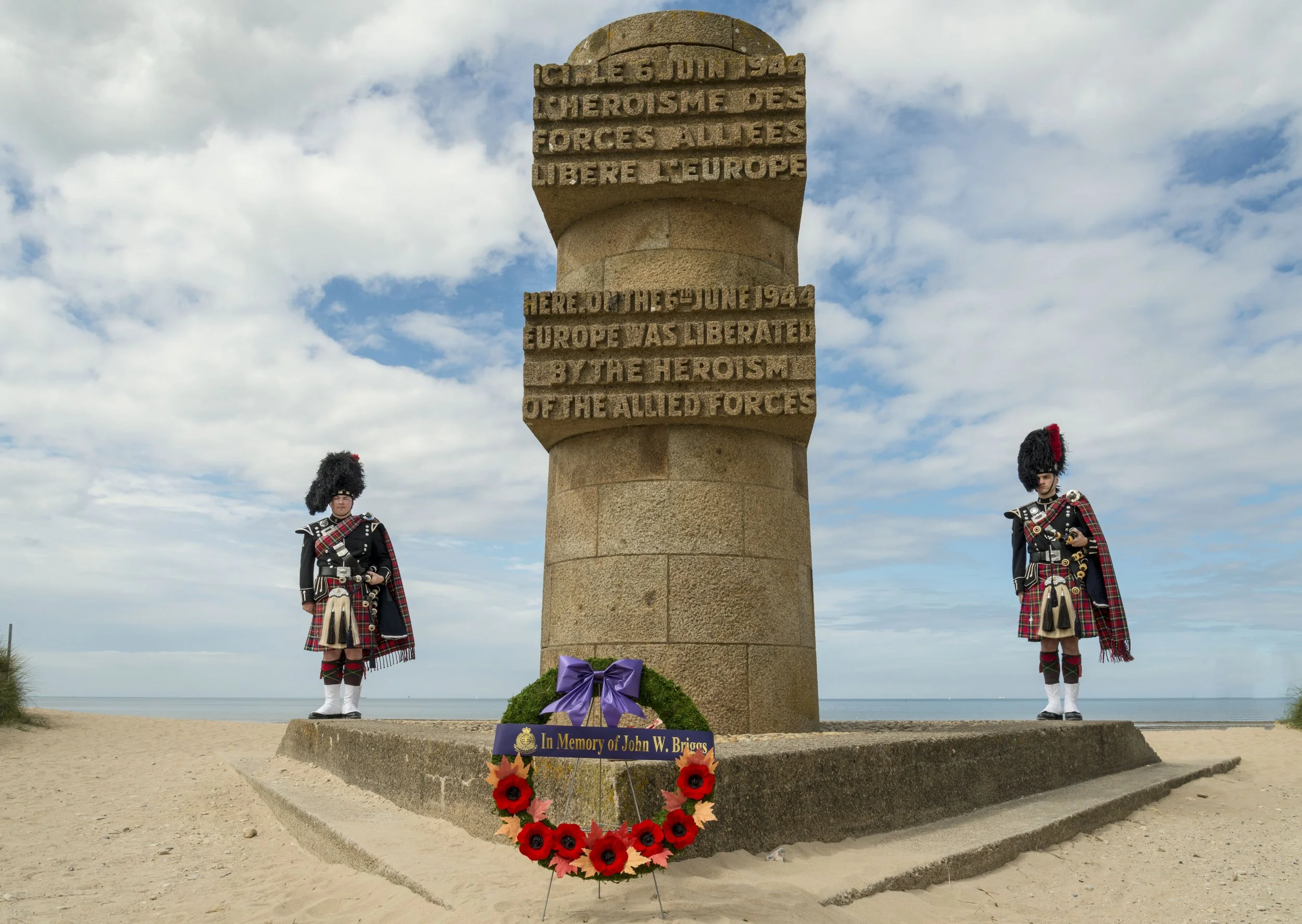 A war memorial on a sandy beach with a large stone pillar inscribed with a dedication to Allied forces. Two soldiers in traditional Scottish kilts and bearskin hats stand guard on either side of the monument. A floral wreath with a purple ribbon and a memorial plaque reading 'In Memory of John W. Briggs' is placed at the base of the monument.
