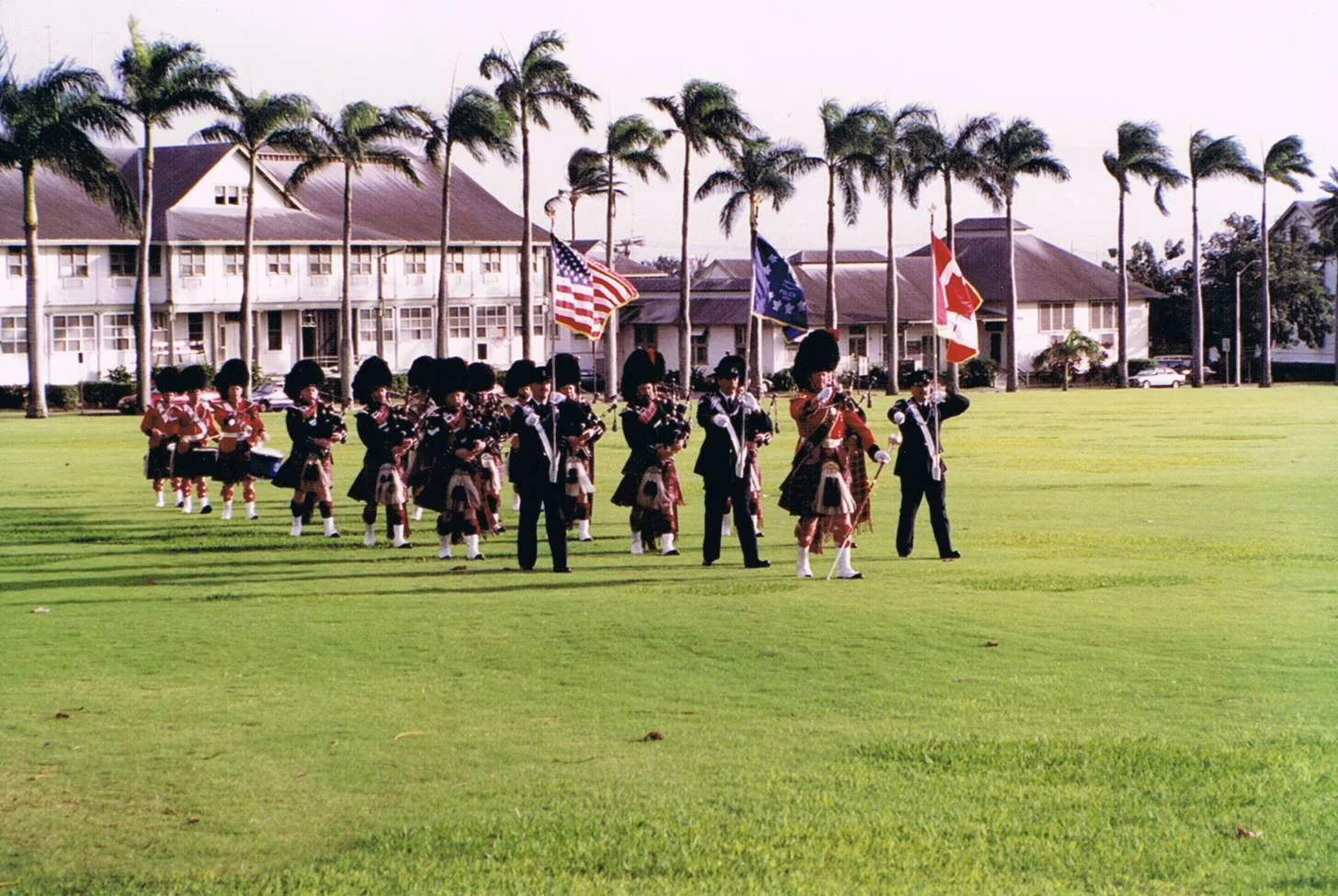 A group of children dressed in Scottish Highland dress, standing in formation on a green lawn, holding flags including the American, Scottish, and Canadian flags, with a row of palm trees and a building with a gabled roof in the background.