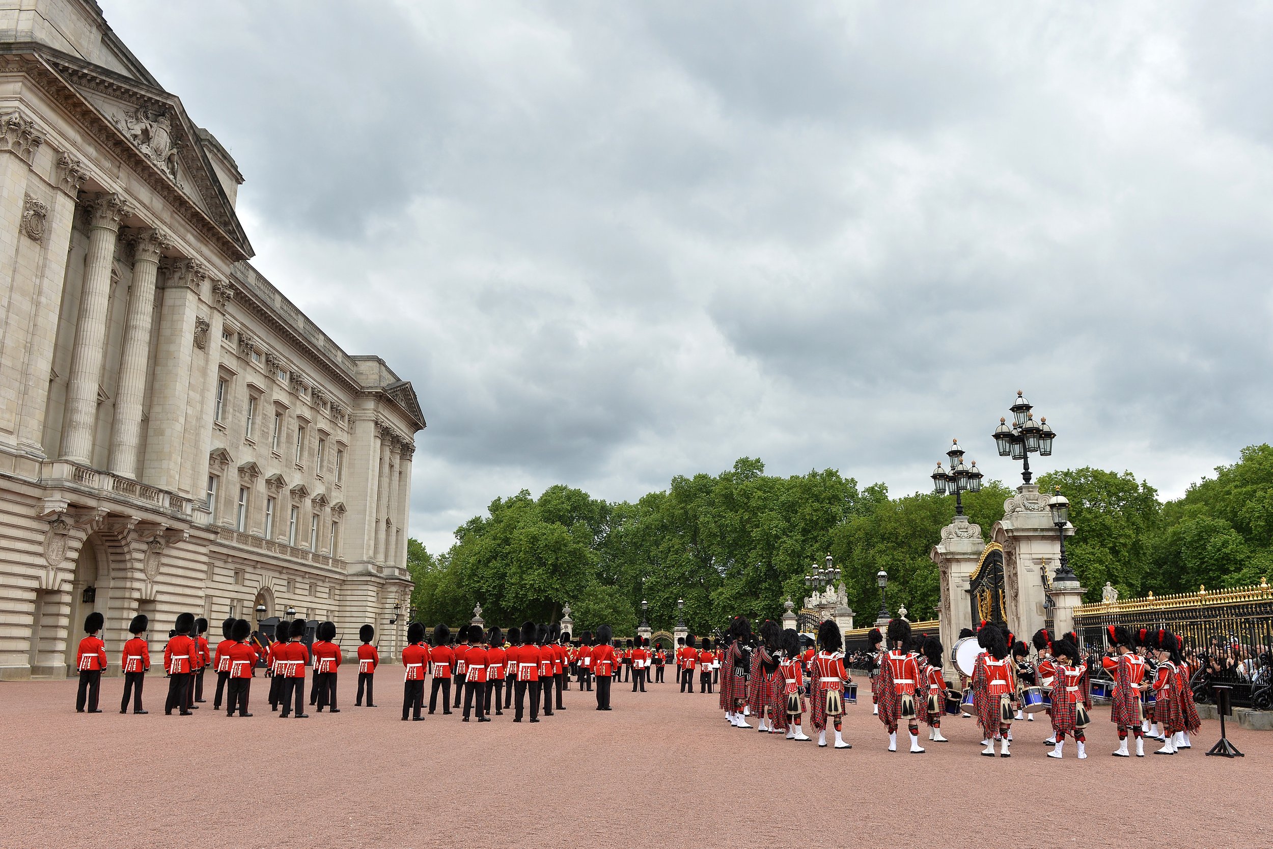 British guards in red uniforms and bearskin hats performing a ceremony outside a large historic building with columns, surrounded by green trees and ornate lamp posts.