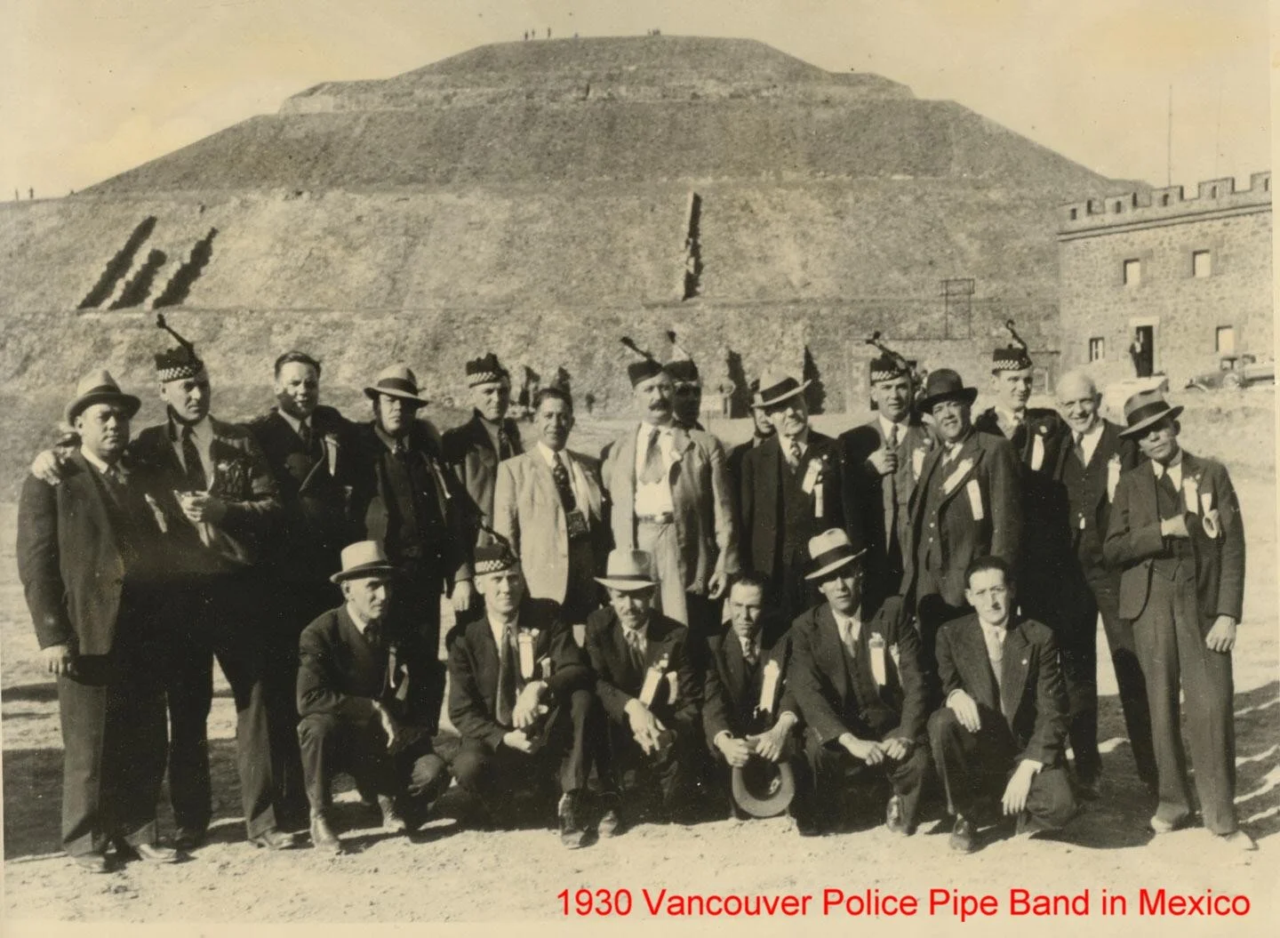A black-and-white photo of the 1930 Vancouver Police Pipe Band in Mexico, with band members in uniforms and hats, standing outdoors in front of a large stepped pyramid and a stone building.