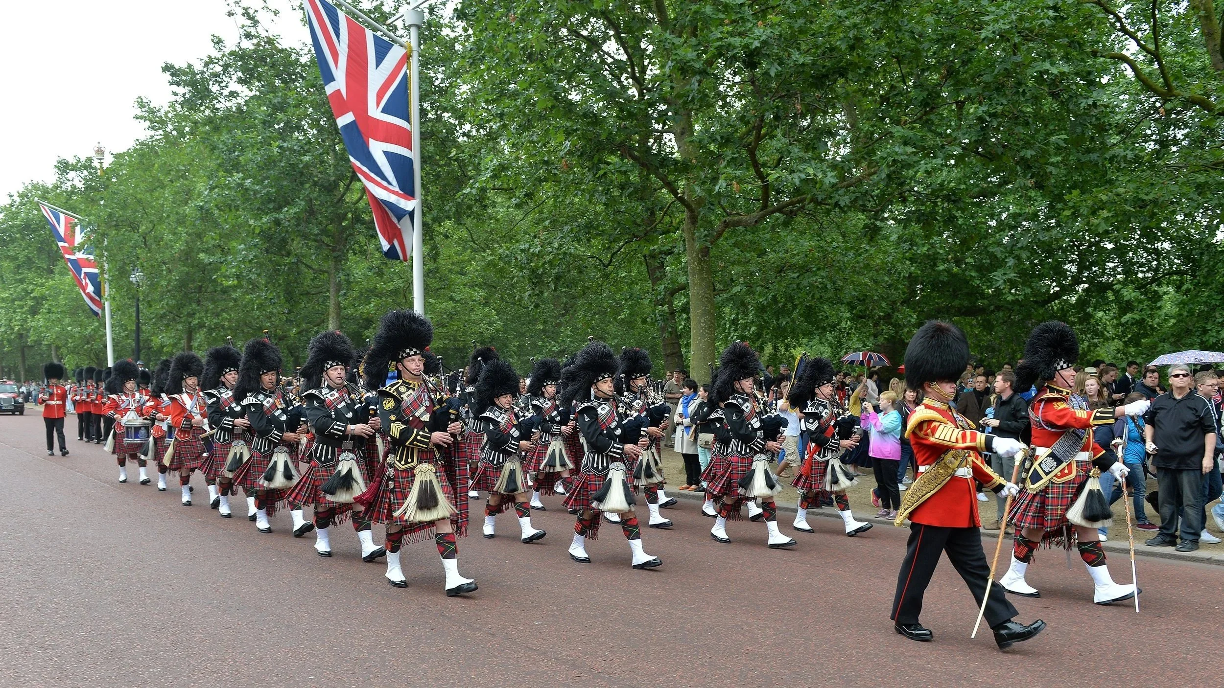 A parade featuring a group of people dressed in traditional Scottish military uniforms, marching in formation along a street with green trees and Union Jack flags flying overhead, while spectators watch on the side.