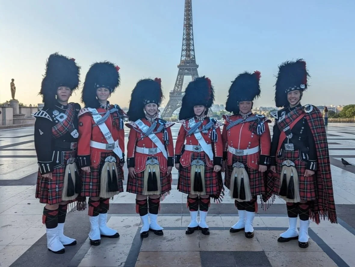 Six women dressed in traditional Scottish Highland dress with large feathered hats, standing in front of the Eiffel Tower in Paris.