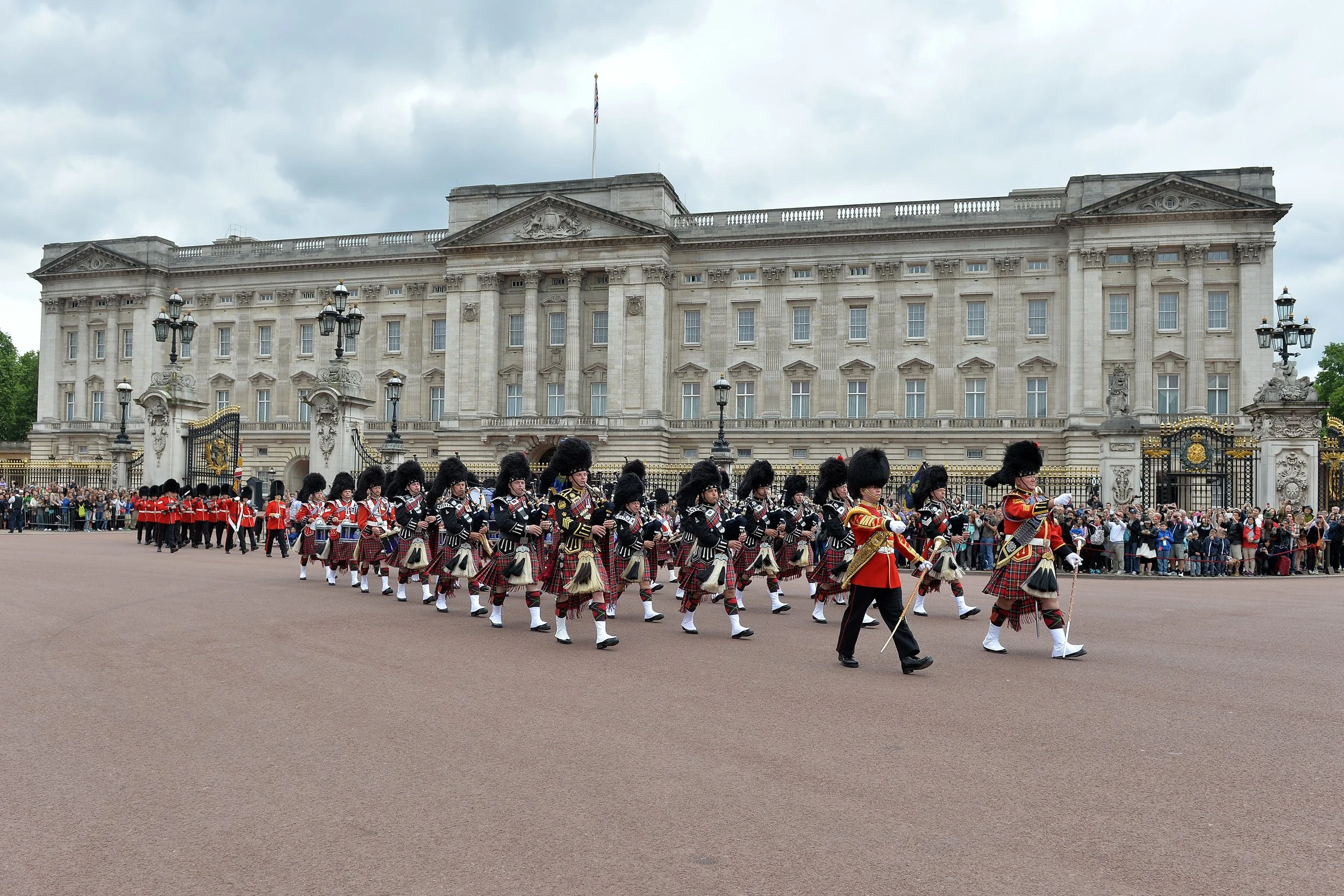 A synchronized march of people dressed in traditional Scottish attire in front of Buckingham Palace, with a gathering of spectators watching.