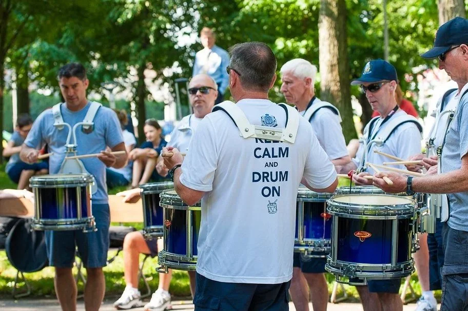 Group of people playing drums outdoors in a park, with trees and onlookers in the background.