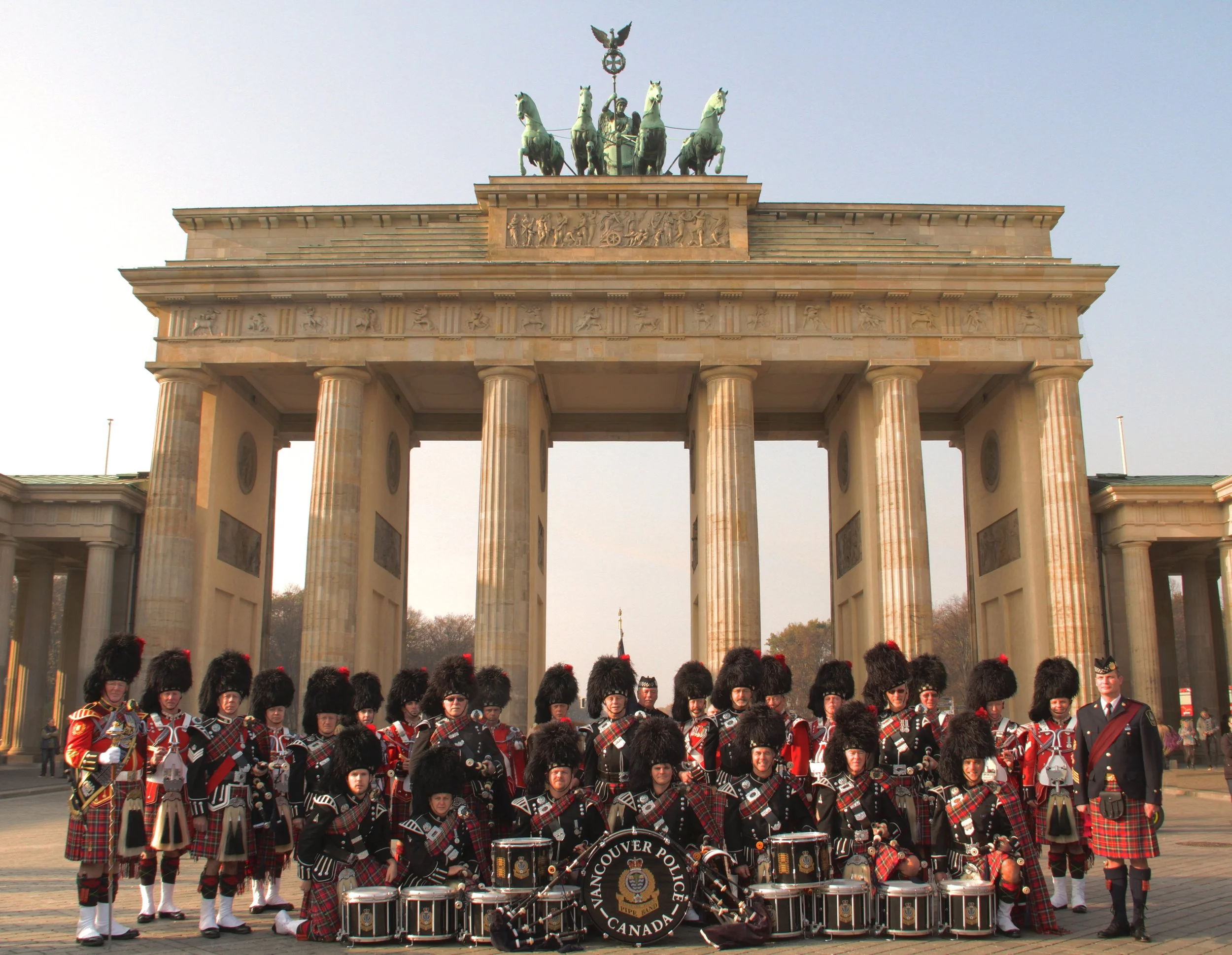 A group of people in traditional Scottish attire, including kilts and bearskin hats, standing in front of the Brandenburg Gate in Berlin, Germany. Some are holding musical instruments and a banner that reads 'Vancouver Police, Canada.'