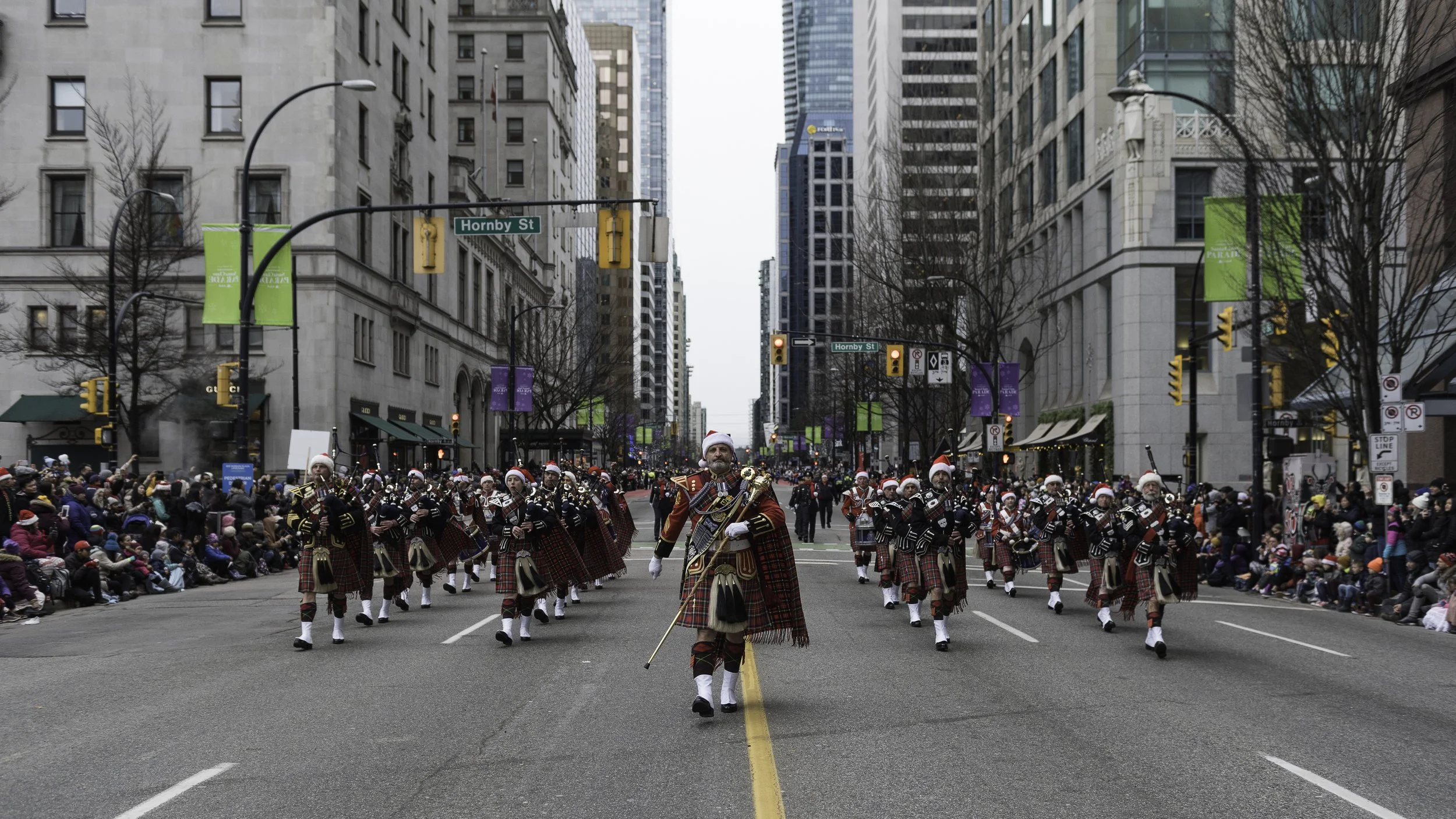 A parade on a city street featuring a marching band wearing Scottish kilts and Santa hats, with spectators seated along the sides of the street, and tall buildings surrounding the scene.