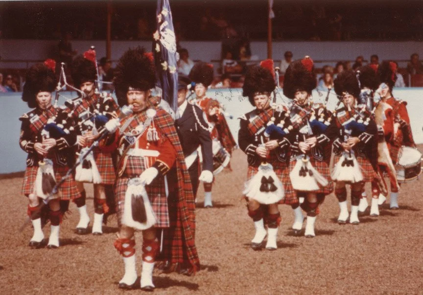 A bagpiper leading a marching band dressed in traditional Scottish kilts and uniforms, performing on a sports field during a parade or event.