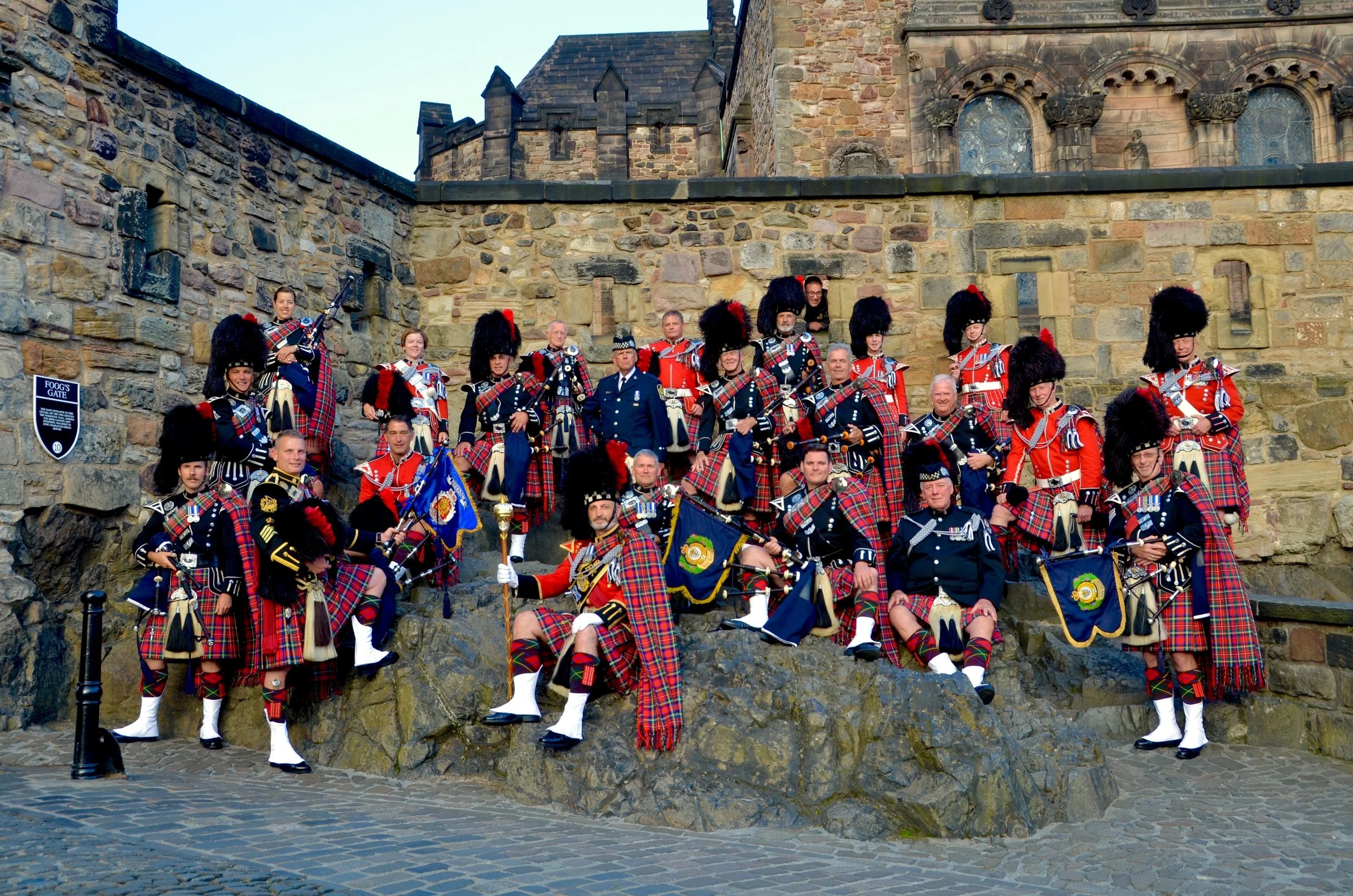A group of people in traditional Scottish kilts and uniforms, some holding bagpipes, posing on a stone steps in front of a historic stone building.