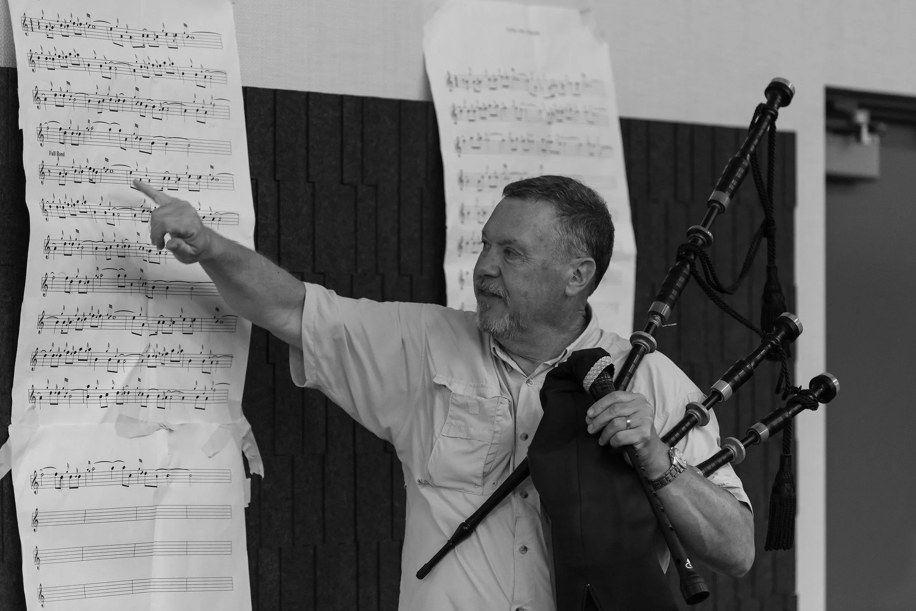 A man holding bagpipes in one hand and a microphone in the other, pointing at sheet music on a wall.