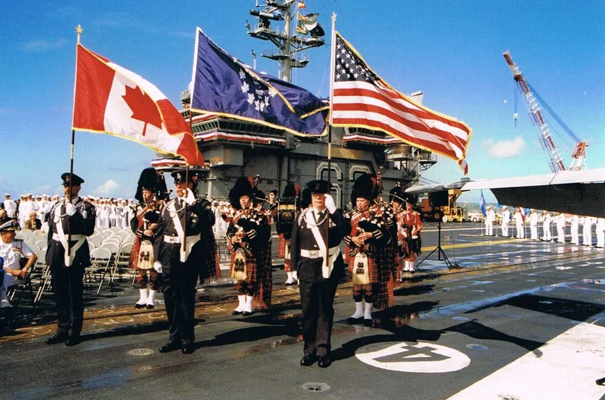 Military personnel in uniform holding flags on the deck of a navy ship, with a crowd of sailors and ceremonial flags including the Canadian, U.S., and a purple naval ensign in the background.