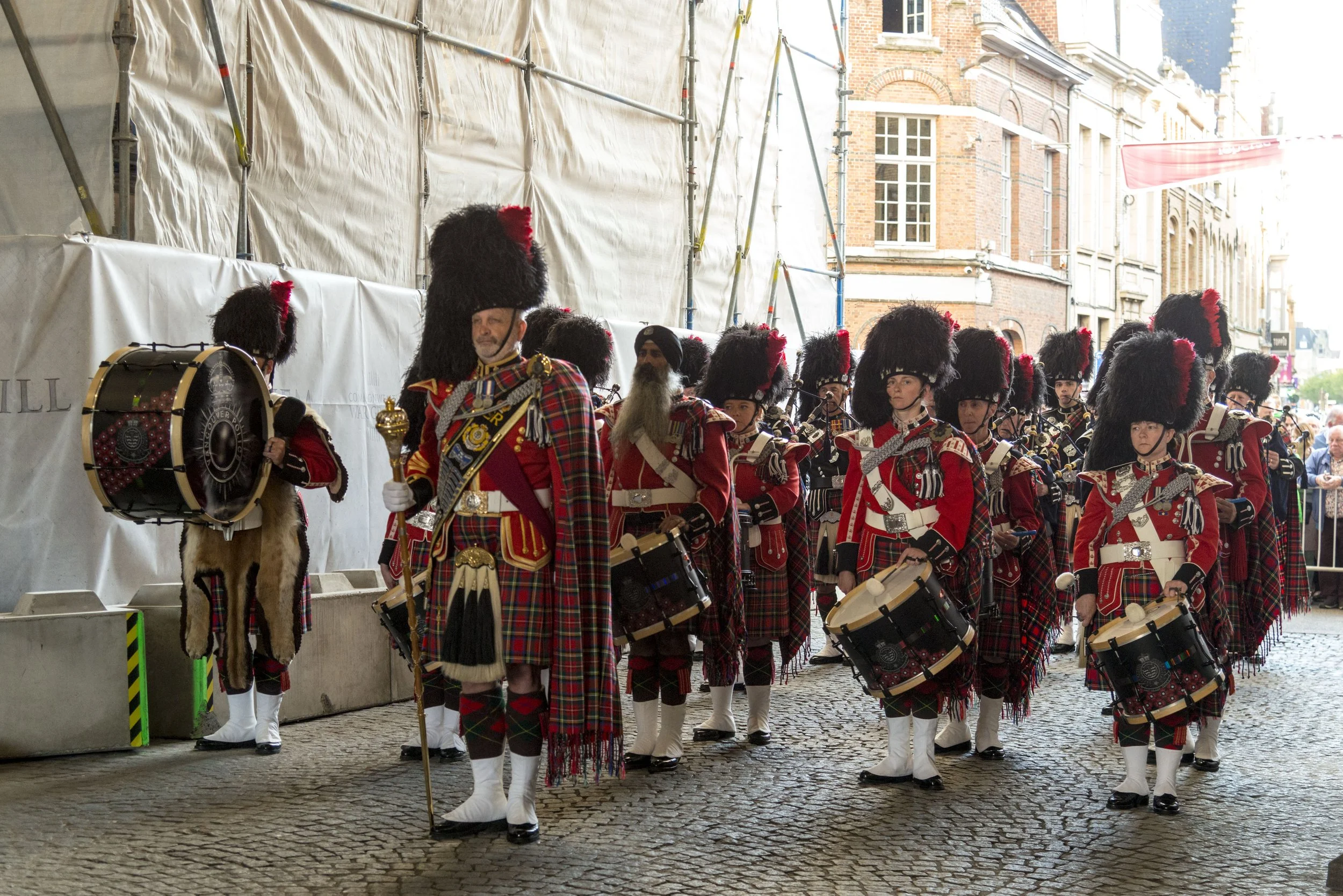 A group of pipers and drummers in traditional Scottish Highland dress, marching in a parade on cobblestone street, with historic brick buildings in the background.
