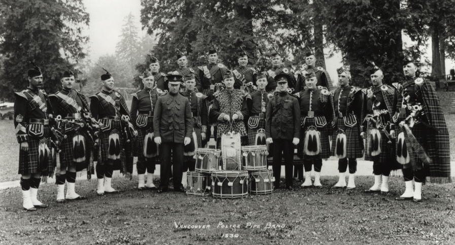 A black-and-white photo of a group of men in Scottish military uniforms, with some holding bagpipes, standing outdoors with trees in the background. They are posing around drums with a person in the center, possibly a leader or conductor. The text at the bottom reads "Vancouver Pipe Band 1930."