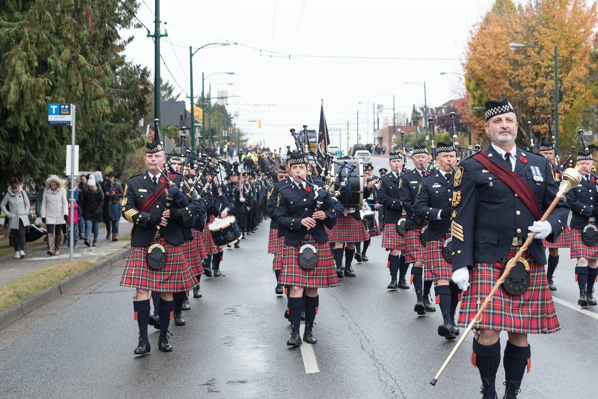 A parade with people dressed in traditional Scottish attire, including kilts, is marching down a street. Spectators are standing on the sidewalk watching the procession.