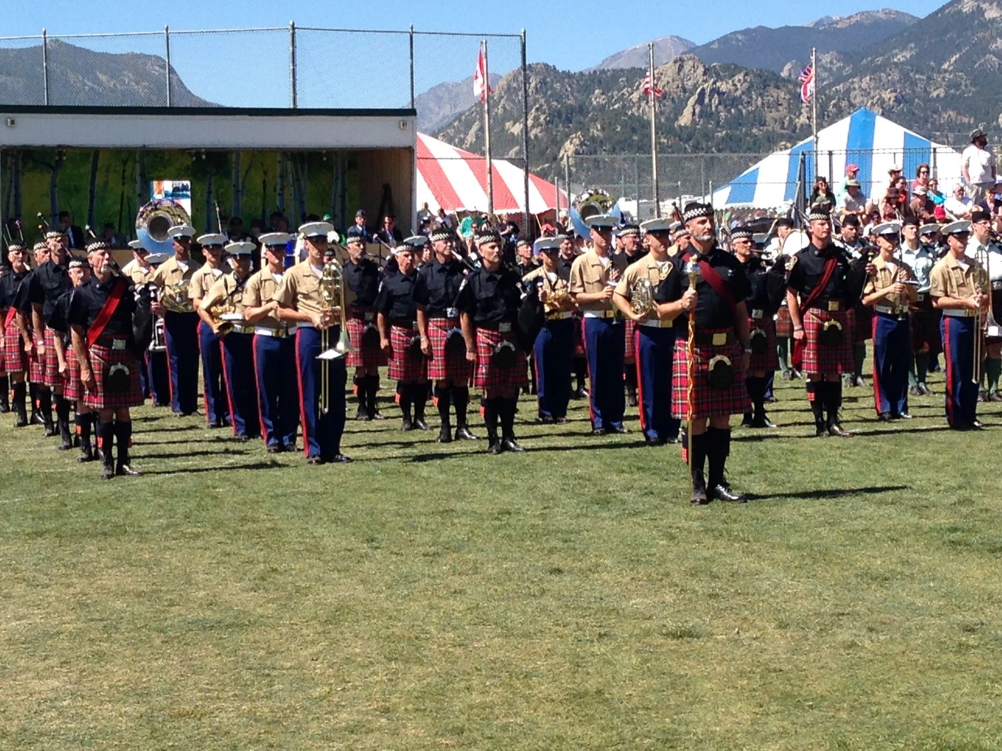 A marching band dressed in uniform, with some members in khaki and others in black, standing on a grassy field during a parade or festival, with tents, flags, and mountains in the background.
