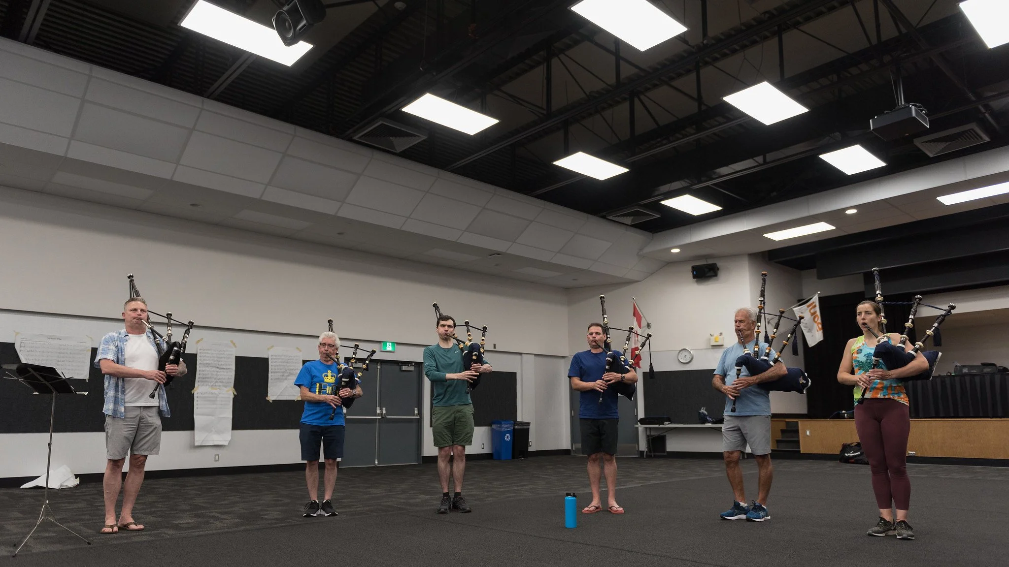 Group of six people standing in a line inside a room, each holding bagpipes. The room has black and white walls, a ceiling with multiple fluorescent lights, and some music sheets taped to the wall.