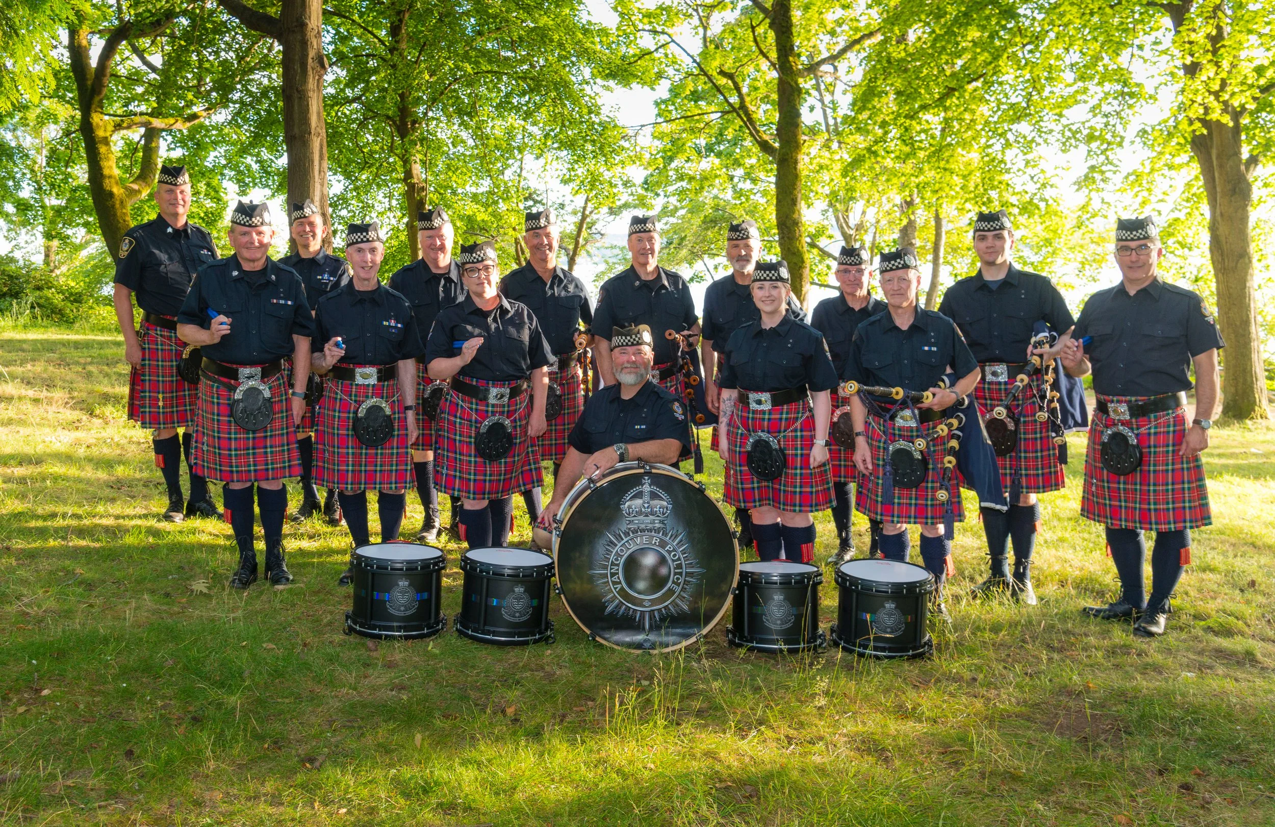 A group of police officers dressed in traditional Scottish kilts and uniforms, standing outdoors among trees, with drums in front of them.
