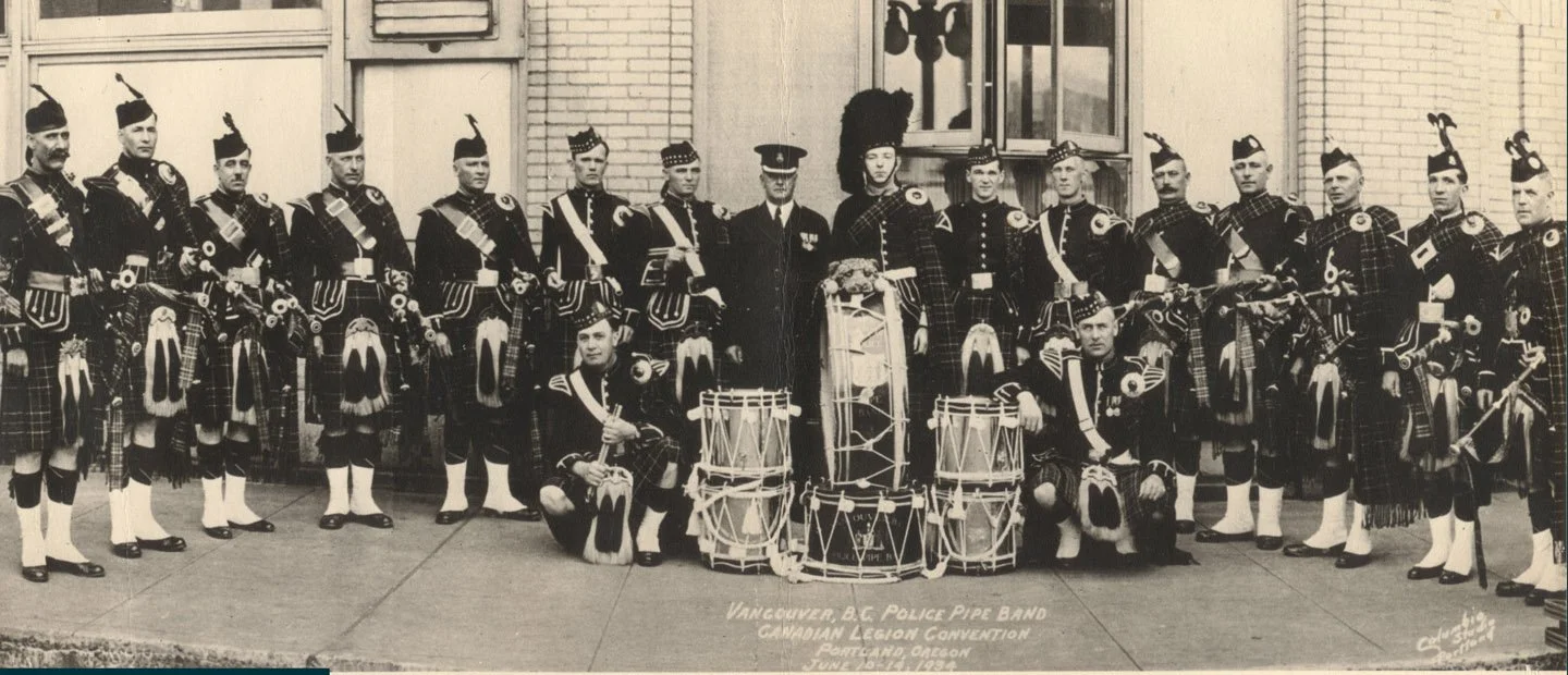 A black and white photo of the Vancouver B.C. Police Pipe Band, with band members dressed in traditional Scottish uniforms, standing in front of a building. There are drums in front and two band members kneeling. The photo is labeled as taken at the Canadian Legion Convention in Portland, Oregon on June 21-22, 1922.