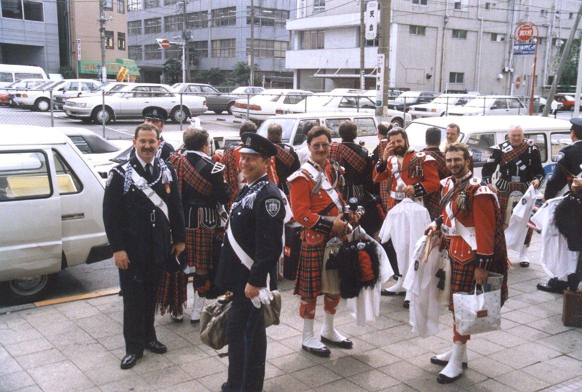 A group of men dressed in traditional Scottish Highland dress, including kilts, jackets, and sporrans, standing outside on a city street. Some are holding bags and others are talking and smiling.