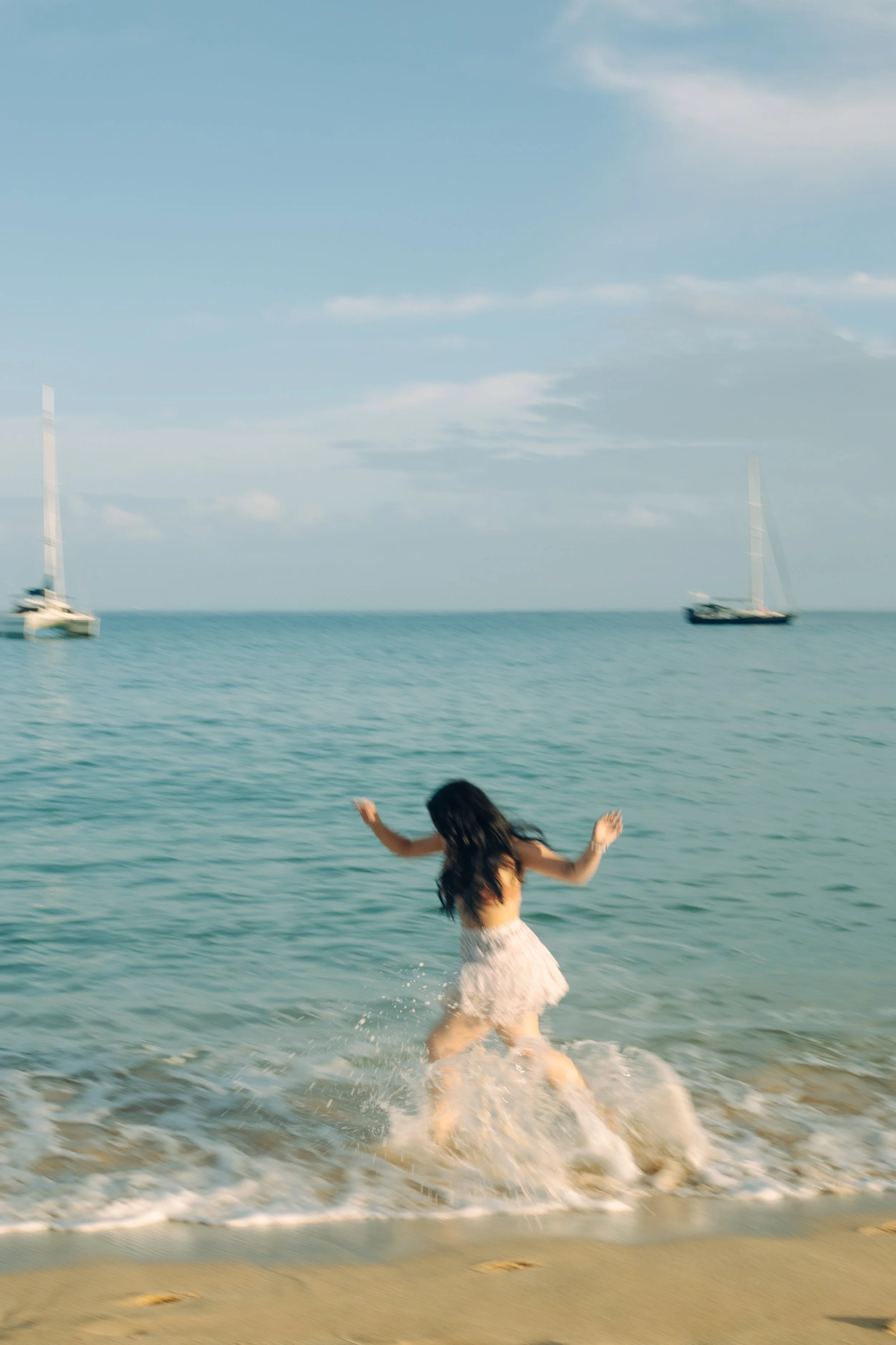A girl playing in shallow water at the beach with sailboats in the background under a blue sky.