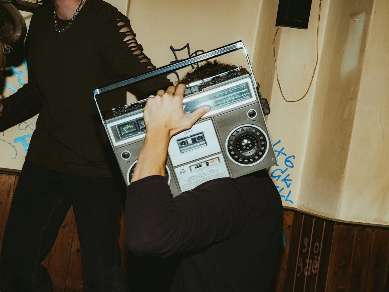Person wearing a boombox on their head, pointing to the radio to a woman wearing a black shirt with cut-out sleeves.