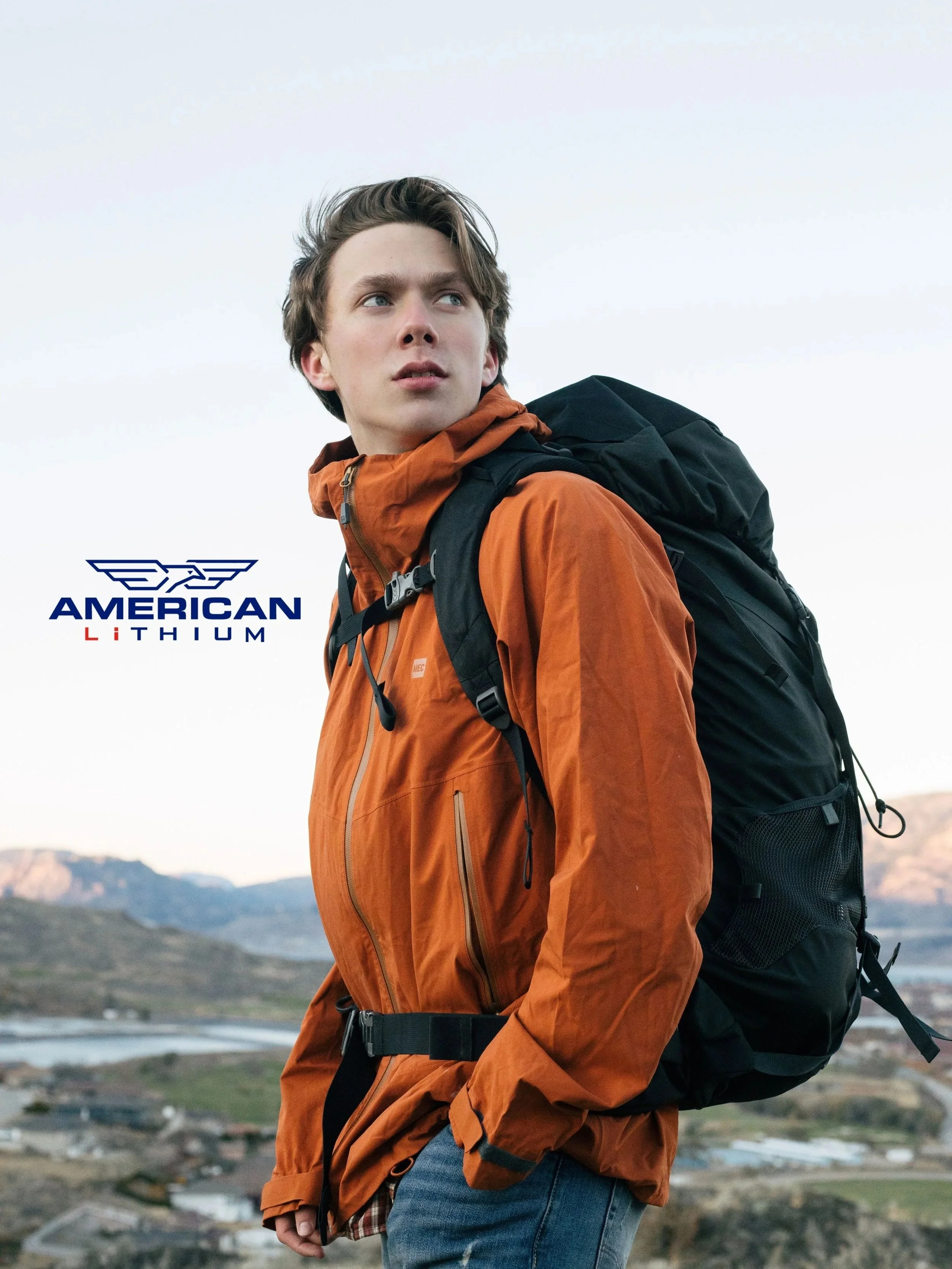 Young man with brown hair wearing an orange jacket and blue jeans carrying a black backpack, standing outdoors with mountains and a river in the background.